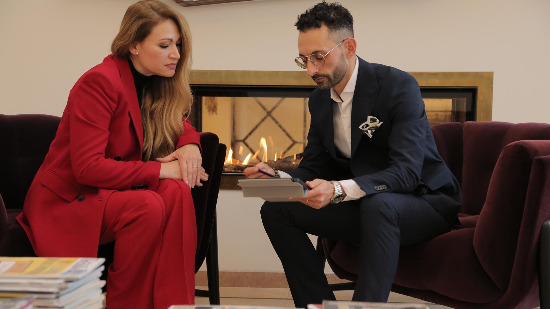 Due persone discutono sedute davanti a un camino, una donna in rosso e un uomo.